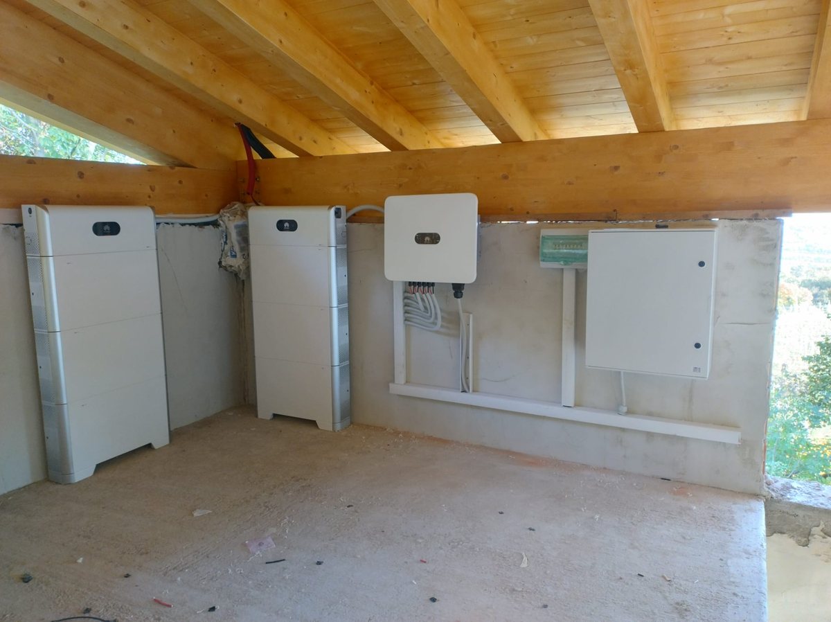 Due batterie bianche impilate, inverter cablato e quadro elettrico su muro di cemento sotto travi in legno